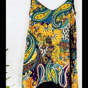 May include: A sleeveless, flowy dress with a paisley print in shades of yellow, blue, green, and purple. The dress has a black ruffle trim at the bottom and a V-neckline. The dress is hanging on a wooden hanger against a white brick wall.