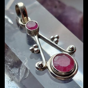 May include: A silver pendant with two round, deep red gemstones. The pendant has a unique, stylized design with a loop at the top for a chain. The Etsy shop URL is visible at the bottom of the image.
