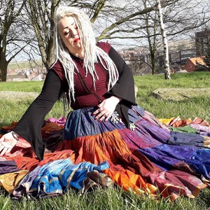 May include: A colorful patchwork skirt with a variety of fabrics in shades of blue, orange, purple, and gold. The skirt is layered and appears to be handmade. The person is wearing a burgundy top with black sleeves.