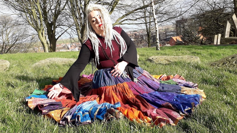 May include: A colorful patchwork skirt with a variety of fabrics in shades of blue, orange, purple, and gold. The skirt is layered and appears to be handmade. The person is wearing a burgundy top with black sleeves.