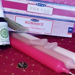 Könnte beinhalten: Eine rosa und weiße Kerze, eine Flasche grüner Flüssigkeit mit einem Herzchakra-Symbol und zwei Schachteln Satya-Räucherstäbchen. Die Räucherstäbchen-Schachteln sind mit "For You" und "Romance" beschriftet.