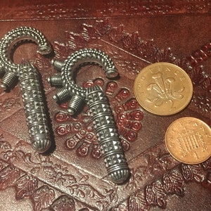 Puede incluir: Dos pendientes plateados ornamentados, con diseño de gancho y detalles geométricos. Se exhiben sobre una superficie marrón rojiza texturizada, junto a una moneda de dos peniques y una de un penique.