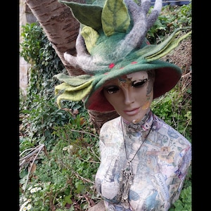 May include: A green felt hat with white and brown antlers and green felt leaves. The hat is on a mannequin with a necklace. The mannequin is standing in front of a tree and green foliage.