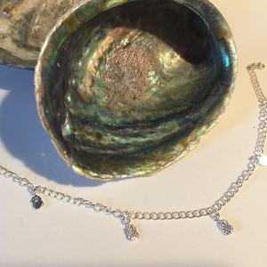 May include: A silver chain bracelet with five silver charms hanging from it. The charms are shaped like hearts and pineapples. The bracelet is lying on a white surface next to a large iridescent seashell.