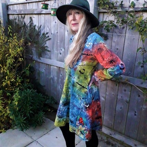 May include: A woman wearing a long-sleeved dress with a tie-dye pattern of colorful mushrooms. The dress has a fitted bodice and a flowing skirt. She is wearing a green hat and has long blonde hair.