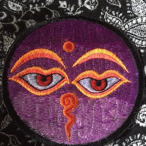 May include: A purple embroidered patch with two eyes, an orange swirl, and a small orange dot in the center. The eyes are white with red pupils and orange outlines.