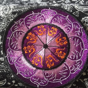 Peut inclure: Patch brodé de couleur violet et orange avec un motif de mandala. Le centre du mandala présente un symbole répété.