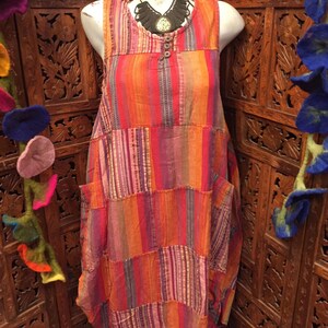 May include: A sleeveless, patchwork dress with a colourful striped pattern in shades of orange, pink, and purple. The dress has a relaxed fit and features two large pockets on the sides.