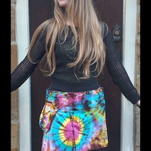 Peut inclure: Une mini-jupe en tie-dye avec une ceinture en cuir marron et une poche zippée. La jupe présente un motif tie-dye coloré avec des nuances de bleu, rose, jaune et marron. La ceinture est ornée de clous métalliques et d'une boucle. La jupe est portée par une personne aux longs cheveux blonds et un pull noir. Le texte "WWW.GUTSYGINGERS.ETSY.COM" est visible en bas de l'image.