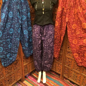 May include: Three pairs of printed pants with a sun and moon design. The pants are in blue, purple, and red. The pants are hanging on a wooden rack.