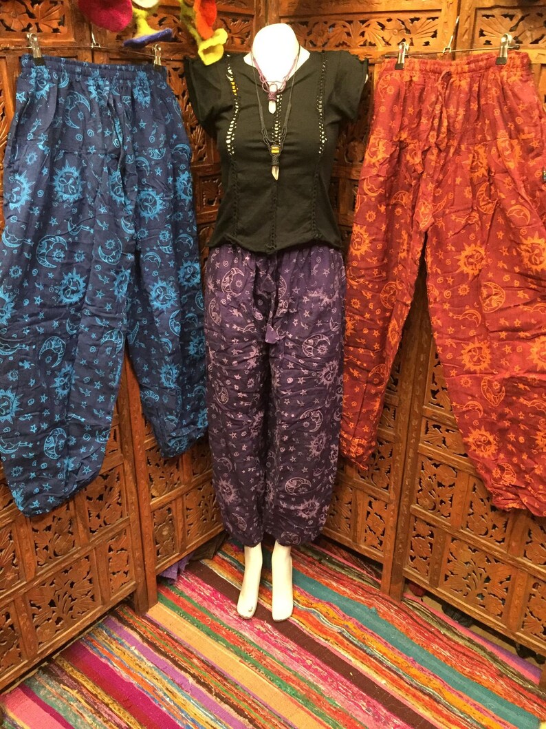 May include: Three pairs of printed pants with a sun and moon design. The pants are in blue, purple, and red. The pants are hanging on a wooden rack.