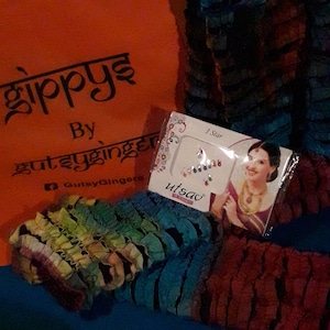 Puede incluir: Un paquete colorido de bindi, o decoraciones para la frente, con una mujer que lleva un sari indio tradicional en la parte delantera. El paquete está etiquetado como "3 Star Utsav" y presenta una variedad de gemas de colores.