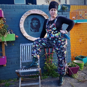 Op de afbeelding: Een vrouw in een zwart shirt en een kleurrijke, gepatroneerde overall staat voor een blauwe bakstenen muur. Ze draagt een zwarte hoofddoek en heeft een groen-blauwe schoonmaakdoek in haar hand. Er is een houten stoel en een verscheidenheid aan potplanten op de achtergrond.