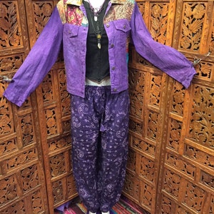 May include: Purple denim jacket with a patchwork collar and a black top underneath. The jacket is worn over a pair of purple pants with a moon and star pattern. The pants are tucked into purple and white slippers with colorful embroidery.