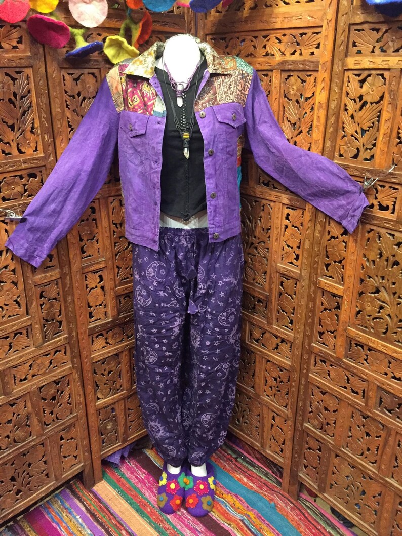 May include: Purple denim jacket with a patchwork collar and a black top underneath. The jacket is worn over a pair of purple pants with a moon and star pattern. The pants are tucked into purple and white slippers with colorful embroidery.