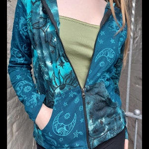 May include: A teal blue hooded sweatshirt with a tie-dye effect and a zip closure. The sweatshirt features a pattern of stars, moons, and suns in a dark blue colour. The sweatshirt has a pocket on the left side.