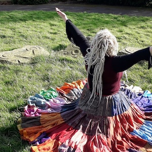 May include: A colorful, tiered skirt with a patchwork design, featuring shades of red, orange, blue, and purple. The skirt is worn with a burgundy top and black sleeves. The person is outdoors on a sunny day, with arms outstretched.