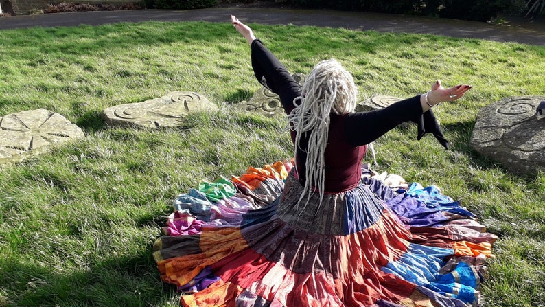 May include: A colorful, tiered skirt with a patchwork design, featuring shades of red, orange, blue, and purple. The skirt is worn with a burgundy top and black sleeves. The person is outdoors on a sunny day, with arms outstretched.