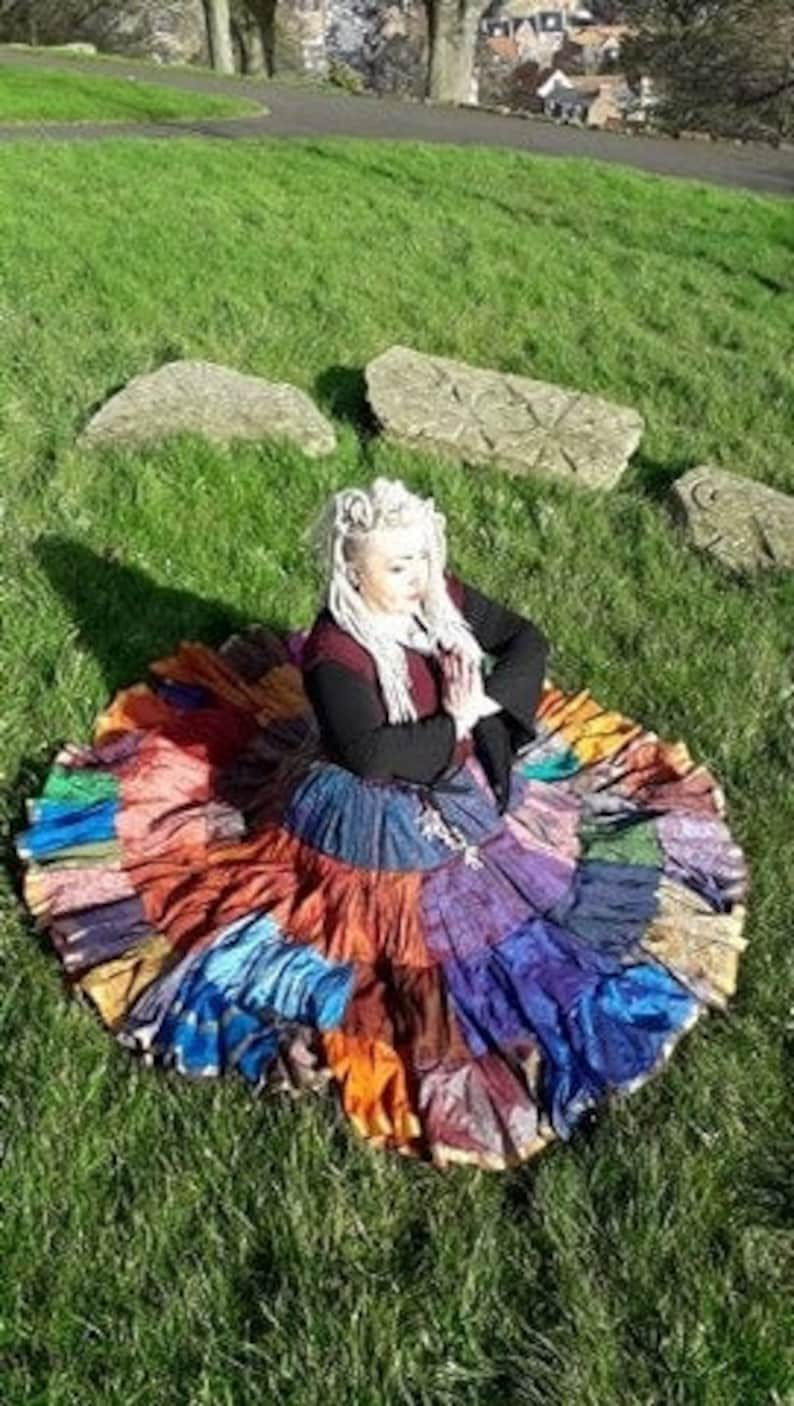 May include: A colorful patchwork skirt with a wide, circular shape. The skirt features a variety of fabrics in shades of blue, orange, purple, and green. The person is wearing a black top and sitting on green grass.