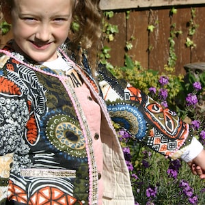 May include: A young girl wearing a colourful, patterned jacket with a pink and white trim. The jacket has a large pocket on the left side. The girl is standing in front of a garden with purple flowers.