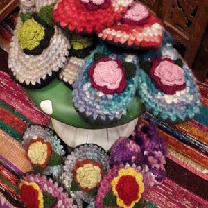Puede incluir: Una colección de zapatillas de crochet de colores con diseños florales. Las zapatillas están hechas con una variedad de colores de hilo, incluyendo rosa, azul, verde y rojo. Las zapatillas están dispuestas sobre una superficie verde con una alfombra a rayas en el fondo.