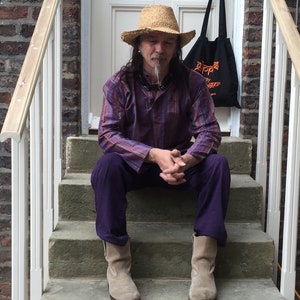 May include: A man wearing a purple plaid shirt, a straw hat, and brown boots sits on a set of stone steps. He is wearing a necklace with a silver pendant. The steps are in front of a white door with a black tote bag hanging on the door handle.