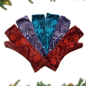 May include: A collection of velvet fingerless gloves in shades of burgundy, teal, and purple, each embroidered with a swirling floral design. The gloves are arranged in a fan shape, showcasing their intricate details and textures. Festive holiday decor surrounds the gloves.