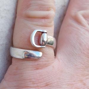 Könnte beinhalten: Ein silberner Ring mit einem Schnallen-Design. Der Ring wird an einem Finger getragen.