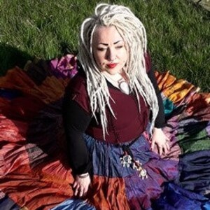 May include: A colorful patchwork skirt with a variety of colors including red, orange, blue, and purple. The skirt is worn with a burgundy top and black long sleeves. The person is sitting on green grass with stone carvings in the background.