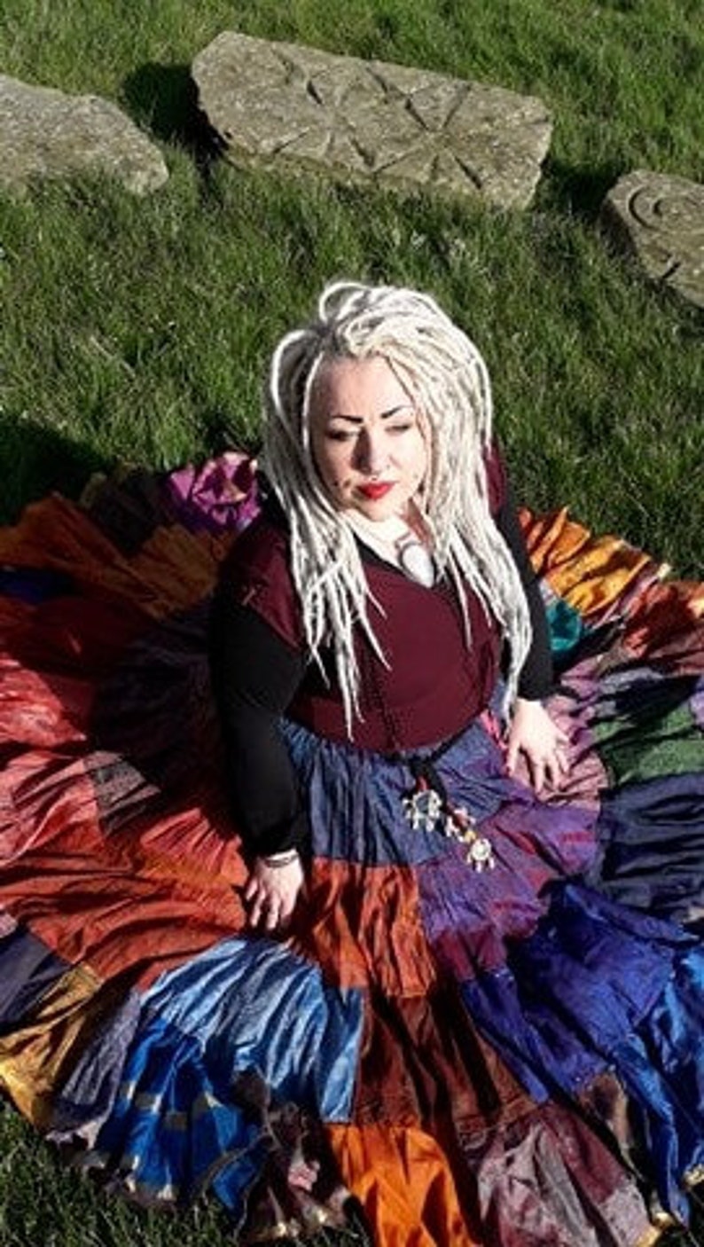 May include: A colorful patchwork skirt with a variety of colors including red, orange, blue, and purple. The skirt is worn with a burgundy top and black long sleeves. The person is sitting on green grass with stone carvings in the background.