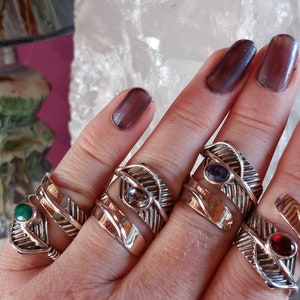 May include: Five silver rings with different colored gemstones set in a feather design. The rings are worn on a hand with dark purple nail polish.