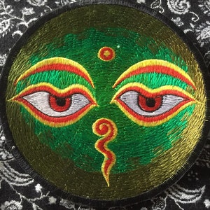 Peut inclure: Un patch rond brodé de couleur verte avec deux yeux rouges, des sourcils jaunes et un tourbillon rouge et jaune au centre. Le patch est entouré d'une bordure noire.