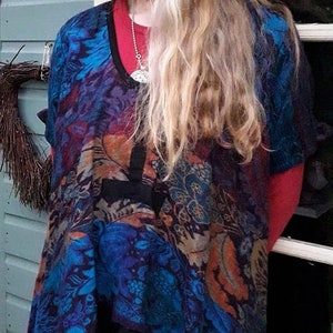 May include: A woman wearing a blue, orange and black patterned tunic with a black undershirt. The tunic has a long, flowing design and is layered over a pair of teal trousers.