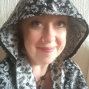 May include: A black and white patterned hooded jacket. The jacket has a floral and paisley design. The hood is up, framing the face. The jacket is open, revealing a necklace.