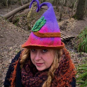 May include: A purple and pink felt witch hat with a green leaf design and a curled brim. The hat has a brown felt band around the base.