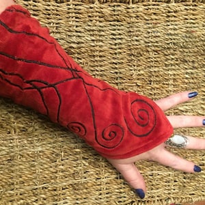 Puede incluir: Puño de brazo de terciopelo rojo con remolinos bordados en negro. El puño se lleva en el brazo izquierdo y se extiende hasta la muñeca.
