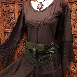 Puede incluir: Una blusa marrón de manga larga con ribete de encaje y una falda a juego. Un cinturón verde con hebilla y detalles de encaje se lleva en la cintura. Se ve un collar con colgante y una piedra marrón.