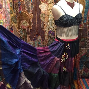 May include: A mannequin displays a bohemian-style outfit. The ensemble includes a beaded black bra top with fringe, a tiered patchwork skirt in shades of purple, blue, and gold, and a matching necklace. The background features a colorful tapestry.
