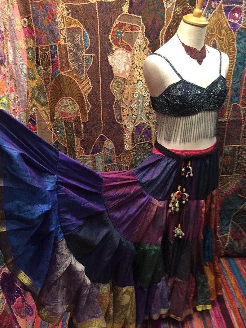 May include: A mannequin displays a bohemian-style outfit. The ensemble includes a beaded black bra top with fringe, a tiered patchwork skirt in shades of purple, blue, and gold, and a matching necklace. The background features a colorful tapestry.