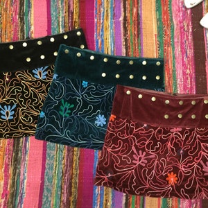 Peut inclure: Trois mini-jupes en velours noir, vert d'eau et bordeaux, chacune avec une bande supérieure contrastante ornée de clous dorés. Les jupes présentent des motifs floraux brodés en or, bleu, vert et rose. Elles sont présentées sur un textile rayé coloré.