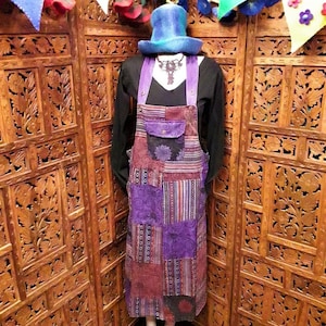 May include: A long purple and brown patchwork overall dress with a large pocket on the front. The dress has a black top and is worn by a mannequin with a blue top hat.