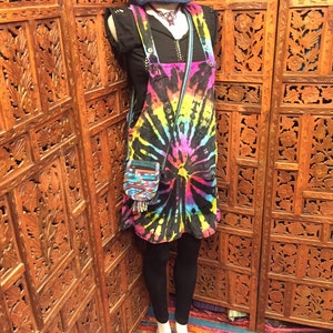 Pode incluir: Um vestido de macac&atilde;o de ganga tie-dye com um top preto e leggings pretos. O vestido tem um padr&atilde;o de tie-dye arco-&iacute;ris com fundo preto. A modelo est&aacute; usando um chap&eacute;u azul e um colar.