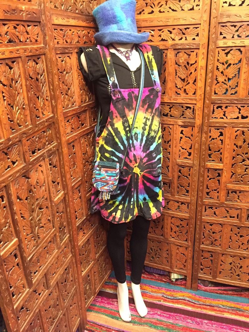 Pode incluir: Um vestido de macac&atilde;o de ganga tie-dye com um top preto e leggings pretos. O vestido tem um padr&atilde;o de tie-dye arco-&iacute;ris com fundo preto. A modelo est&aacute; usando um chap&eacute;u azul e um colar.