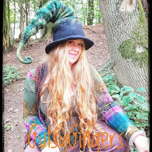 May include: A woman wearing a black and green felt witch hat with a long, curled green brim. The hat is decorated with a green and blue swirl pattern. She is wearing a colourful, tie-dye shirt and has long, blonde hair.