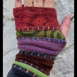 Peut inclure: Manchette de poignet multicolore composée de couches de tissu bordeaux, vert olive, violet, marron et noir. La manchette est ornée de coutures décoratives de différentes couleurs. L'adresse du site web est visible en bas.