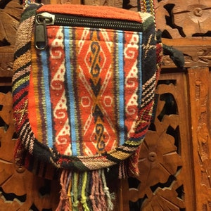 Pode incluir: Uma pequena bolsa tecida colorida com fecho de correr e uma franja longa. A bolsa tem um padr&atilde;o geom&eacute;trico em tons de vermelho, azul e branco. A bolsa est&aacute; pendurada numa superf&iacute;cie de madeira.