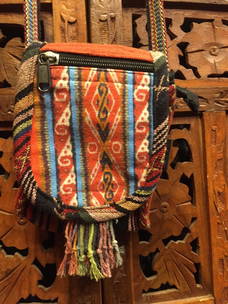 Pode incluir: Uma pequena bolsa tecida colorida com fecho de correr e uma franja longa. A bolsa tem um padr&atilde;o geom&eacute;trico em tons de vermelho, azul e branco. A bolsa est&aacute; pendurada numa superf&iacute;cie de madeira.