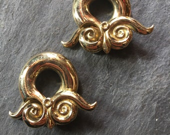 May include: Two gold-colored metal ear gauges with a swirling design. The gauges are shaped like a ring with a decorative swirl on each side.
