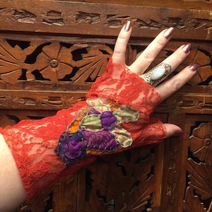 Può includere: Un guanto senza dita in pizzo rosso con un'applicazione in tessuto colorato. Il guante è indossato su una mano con un anello d'argento con una pietra bianca.