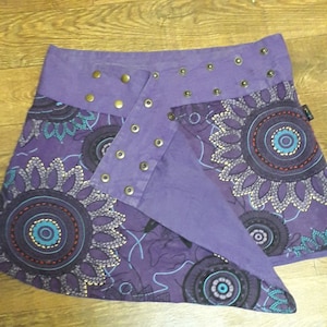 Peut inclure: Jupe violette avec un design enveloppant et une ceinture décorative avec des œillets métalliques. La jupe présente un motif floral coloré avec des cercles et des volutes.